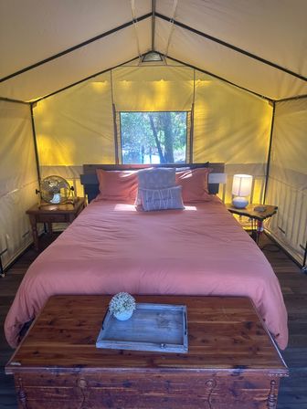 Sunlit glamping tent interior with coral bedding on a king-size bed, rustic wooden chest and tray at the foot, bedside tables with lamp and fan, and a window framing trees and a lake view.