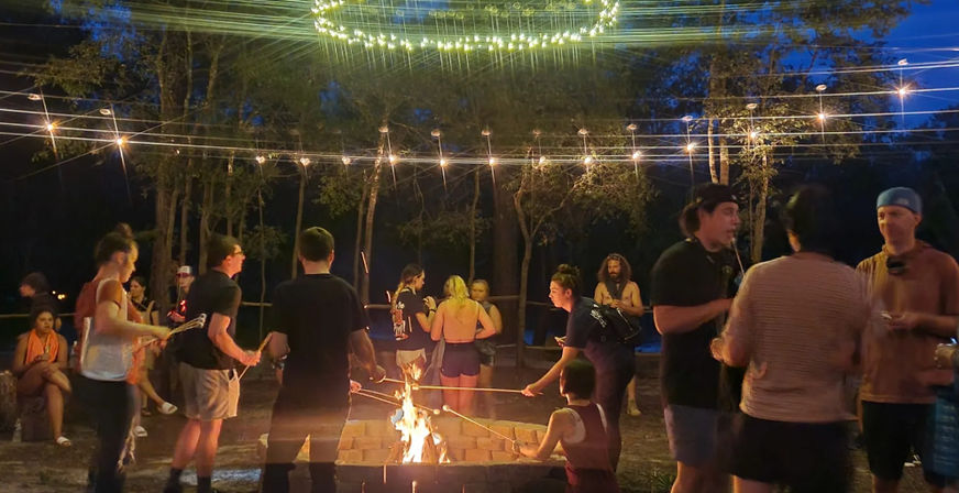 Outdoor nighttime campfire gathering in a wooded area, people roasting marshmallows over a square fire pit under string lights and a circular light fixture, casual summer clothing and relaxed festival vibe.
