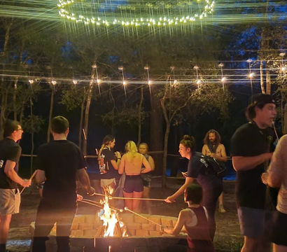 Outdoor nighttime campfire gathering in a wooded area, people roasting marshmallows over a square fire pit under string lights and a circular light fixture, casual summer clothing and relaxed festival vibe.
