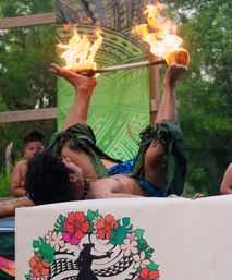 Polynesian fire performer lying on their back on stage, balancing a flaming torch between raised feet, wearing leaf anklets and blue sarong against a tropical patterned backdrop with onlookers nearby.