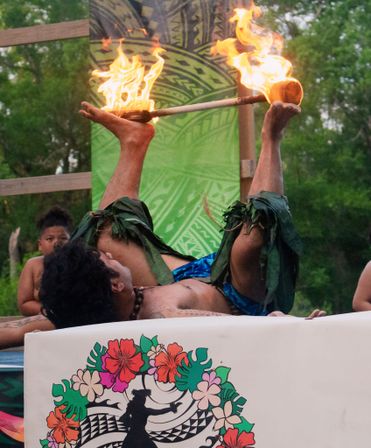 Polynesian fire performer lying on their back on stage, balancing a flaming torch between raised feet, wearing leaf anklets and blue sarong against a tropical patterned backdrop with onlookers nearby.