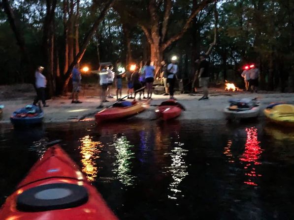 Dusk kayaking scene: red kayak bow in foreground with a group gathered on a tree-lined sandy riverbank, multiple kayaks on shore, a small campfire and colorful light reflections on the water.