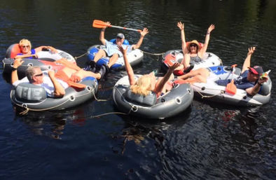 Six people lounging in linked inflatable tubes, cheering and waving orange paddles while floating together on calm summer river water.
