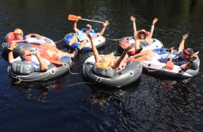 Six people lounging in linked inflatable tubes, cheering and waving orange paddles while floating together on calm summer river water.
