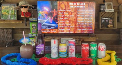 Wood-paneled island visitor center counter with TV showing an adventure activity menu, a mannequin head with sun hat and sunglasses, rows of bottled and canned drinks, colorful leis and a coconut-shaped cocktail cup with flower straw.