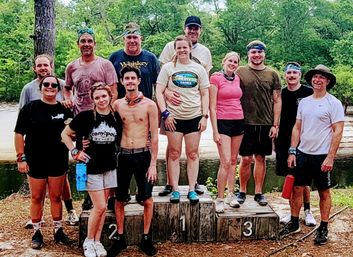 12-person group on a wooden winners' podium by a river in a leafy forest — smiling in activewear and headbands after an outdoor team-building challenge.
