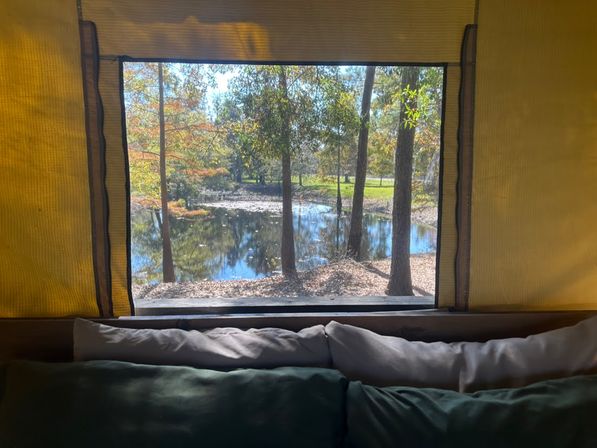 Cozy camping tent window framing a reflective pond and autumn trees, cushions on the windowsill creating a relaxing nature view.