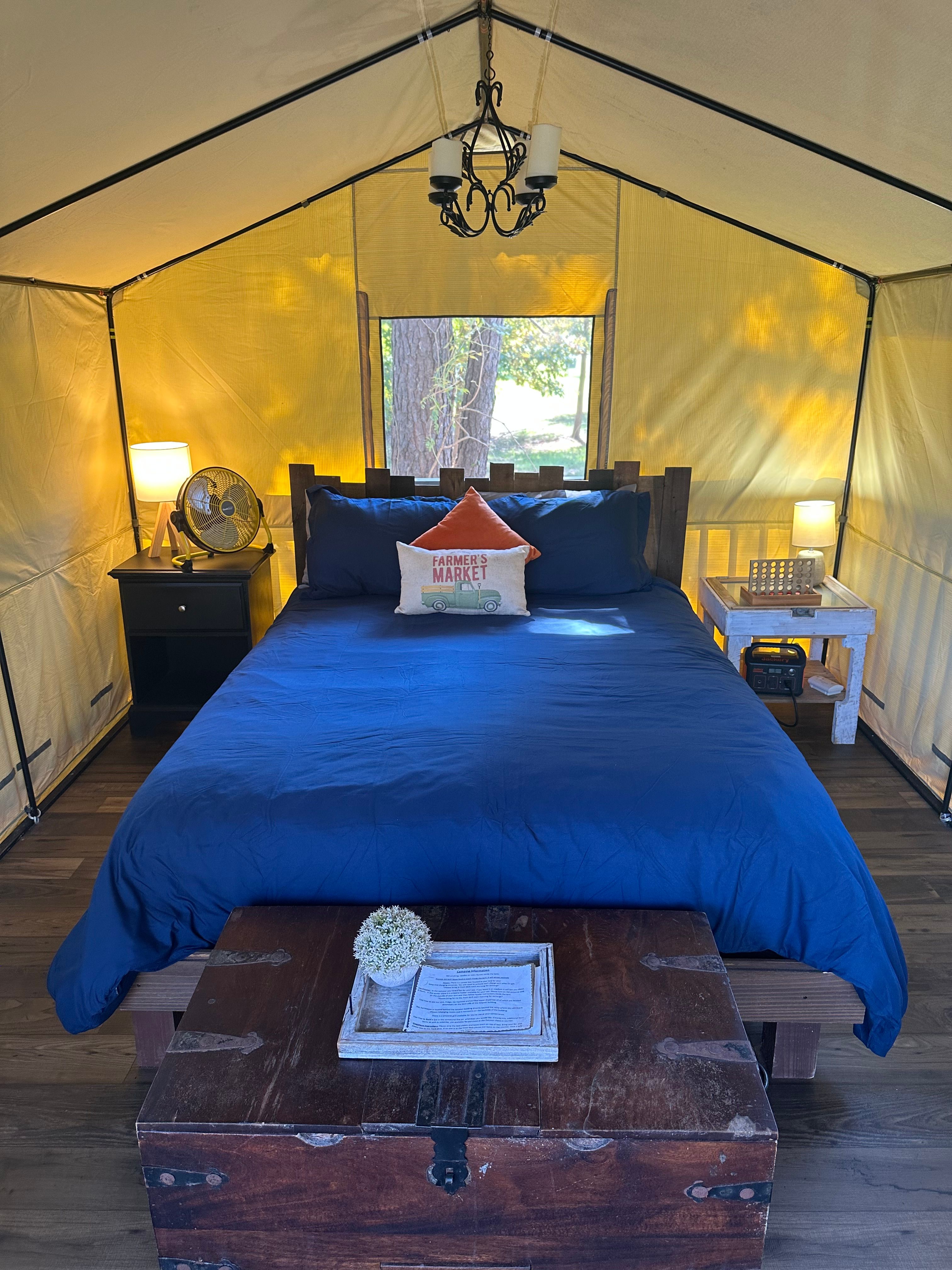 Cozy glamping tent bedroom with navy bedding and wooden headboard, farmhouse "Farmer's Market" accent pillow, bedside lamps, fan, chandelier, vintage trunk at foot and small window framing trees