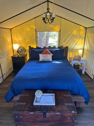 Cozy glamping tent bedroom with navy bedding and wooden headboard, farmhouse "Farmer's Market" accent pillow, bedside lamps, fan, chandelier, vintage trunk at foot and small window framing trees