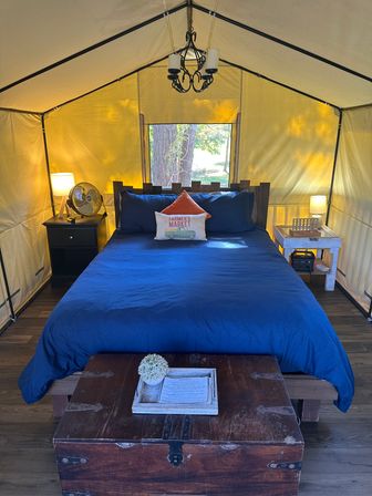 Cozy glamping tent bedroom with navy bedding and wooden headboard, farmhouse "Farmer's Market" accent pillow, bedside lamps, fan, chandelier, vintage trunk at foot and small window framing trees