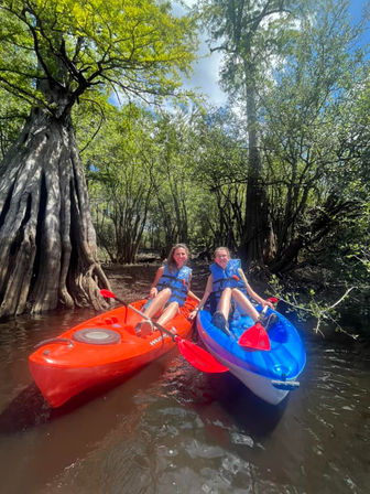 Two kayakers in red and blue kayaks wearing life jackets paddle calm brown water through a sunlit cypress swamp with towering buttressed trees and green canopy