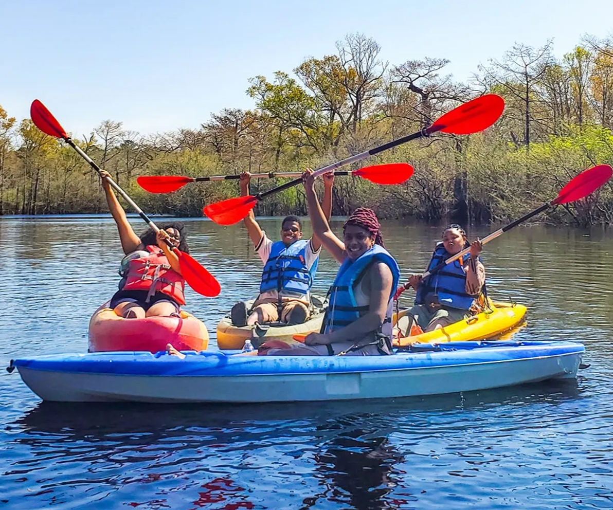 Four adults in life jackets kayaking on a calm lake in colorful kayaks (blue, yellow, red), holding red paddles overhead with spring trees and a sunny sky in the background.
