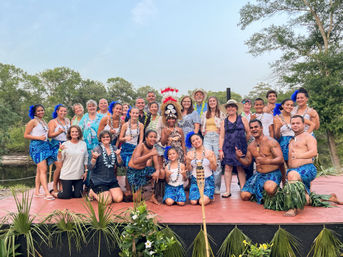 Festive luau group photo of Polynesian dancers and guests in blue sarongs and leis posing on an outdoor riverside stage with palms and trees in the background.