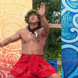 Shirtless dancer wearing a leafy head crown, black bead necklace with crescent pendant and red patterned sarong performing a traditional island dance on a colorful outdoor festival stage.