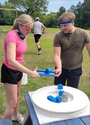 Two players in a sunny park balancing bright blue wooden blocks on a round white target during an outdoor team-building challenge on a grassy field.