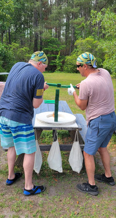 Two men wearing colorful bandanas adjust a green wooden balancing game on a picnic table with canvas weight bags in a grassy, wooded park