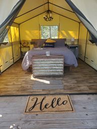 Cozy glamping tent interior with a made bed in gray linens and welcome pillow, rustic trunk at the foot, wood-look floors, bedside tables and fan, yellow canvas walls and chandelier, and a 'Hello' doormat at the entrance.
