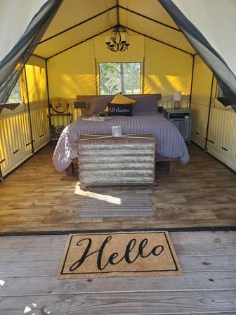 Cozy glamping tent interior with a made bed in gray linens and welcome pillow, rustic trunk at the foot, wood-look floors, bedside tables and fan, yellow canvas walls and chandelier, and a 'Hello' doormat at the entrance.