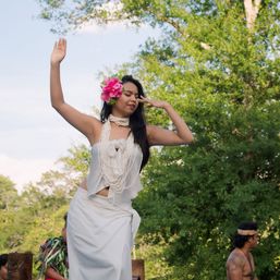 Hula dancer outdoors in white traditional dress with layered shell leis and a bright pink hibiscus in her hair, gracefully posing with arms raised against lush green trees.