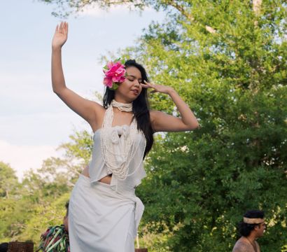 Hula dancer outdoors in white traditional dress with layered shell leis and a bright pink hibiscus in her hair, gracefully posing with arms raised against lush green trees.