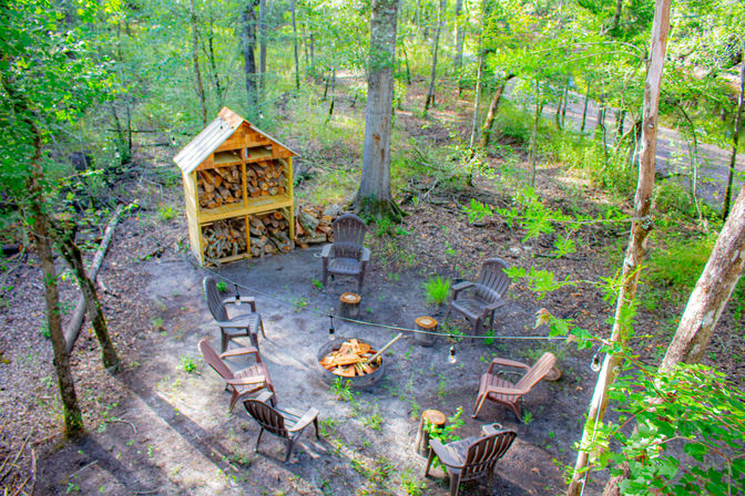 Cozy woodland fire-pit circle in a forest clearing with Adirondack-style chairs around a metal fire pit, stacked firewood in a wooden shelter, string lights overhead, and trees with leafy green undergrowth.