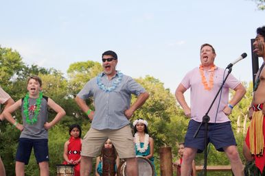 Outdoor summer festival: two men and a boy wearing leis strike playful warrior poses with tongues out during a Pacific Island dance, backed by drummers and costumed dancers.