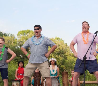 Outdoor summer festival: two men and a boy wearing leis strike playful warrior poses with tongues out during a Pacific Island dance, backed by drummers and costumed dancers.