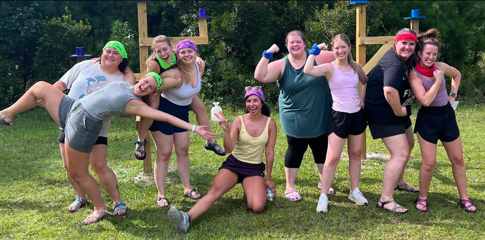 Smiling group of nine people in colorful bandanas and casual athletic wear striking playful poses on a grassy field in front of a wooden outdoor course, team-building picnic vibe