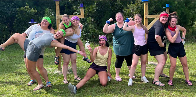 Smiling group of nine people in colorful bandanas and casual athletic wear striking playful poses on a grassy field in front of a wooden outdoor course, team-building picnic vibe