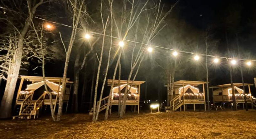 Cozy elevated wooden glamping cabins on stilts in a leafless woodland, twinkling string lights and fairy lights illuminating a nighttime campground.