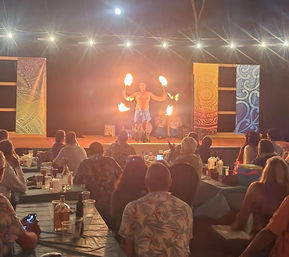 Lively shirtless fire dancer twirling flaming torches on a colorful stage under string lights as a seated audience in tropical shirts watches from tables.