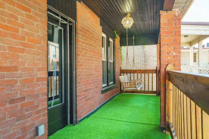 Covered residential porch with bright green artificial turf, red brick walls, wooden railing and a hanging wooden swing under a woven pendant light.