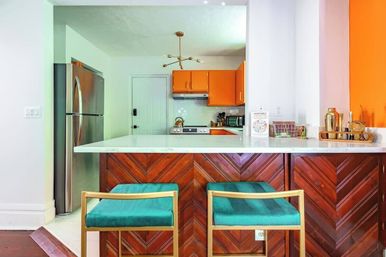 Open-plan kitchen with white marble countertop island and chevron wood paneling, two teal velvet bar stools with gold frames, bright orange cabinets, stainless refrigerator, brass sputnik chandelier and countertop barware.