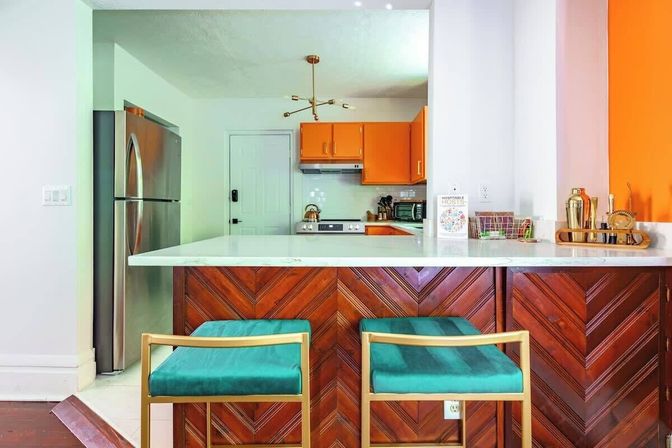 Open-plan kitchen with white marble countertop island and chevron wood paneling, two teal velvet bar stools with gold frames, bright orange cabinets, stainless refrigerator, brass sputnik chandelier and countertop barware.