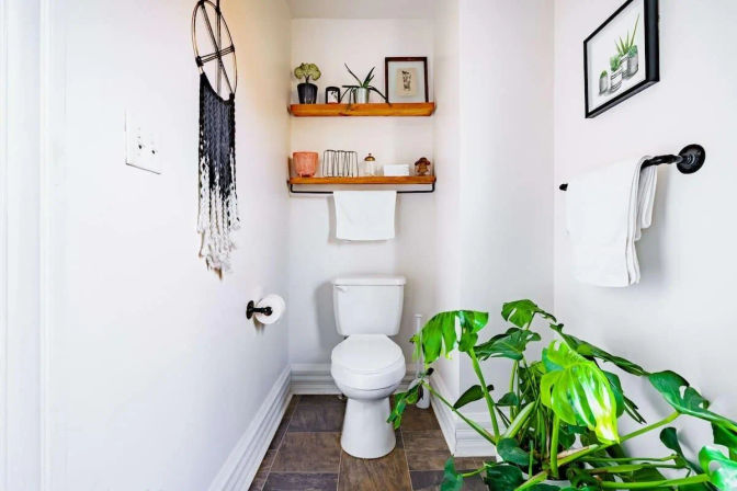 Bright modern powder room with white toilet, wooden floating shelves styled with small plants and decor, macramé wall hanging, white towels on black hardware, and a large potted green plant for a fresh, minimalist vibe.