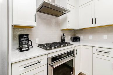 Sleek modern white kitchen corner with stainless steel gas cooktop and oven, range hood, subway tile backsplash, marble countertops, black coffee maker, toaster and knife block