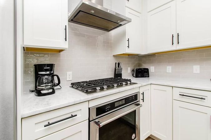 Sleek modern white kitchen corner with stainless steel gas cooktop and oven, range hood, subway tile backsplash, marble countertops, black coffee maker, toaster and knife block