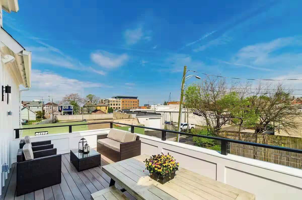 Sunny urban rooftop deck with wicker sofa and chairs, coffee table, picnic-style dining table with potted flowers, glass railing and city building views under a bright blue sky.
