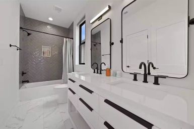 Bright modern white bathroom with double vanity and black matte faucets, gray herringbone-tile tub/shower, and marble-look floor.