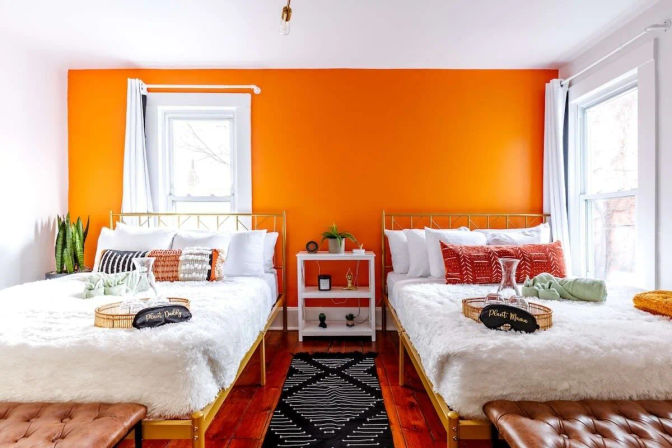 Cheerful twin bedroom with vibrant orange accent wall, gold metal bed frames, fluffy white faux-fur bedding, patterned throw pillows, potted plants, hardwood floors and bright natural light from two windows.