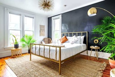 Sunlit boho bedroom with a gold metal bed frame and white bedding against a black accent wall, rattan chair, large indoor plants, jute area rug on hardwood floors, and brass arc floor lamp.