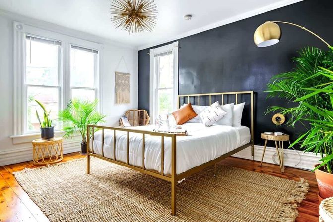 Sunlit boho bedroom with a gold metal bed frame and white bedding against a black accent wall, rattan chair, large indoor plants, jute area rug on hardwood floors, and brass arc floor lamp.