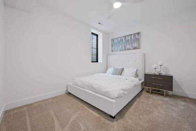 Bright modern minimalist bedroom interior with white upholstered platform bed and white bedding, beige carpet, small vertical window, gray two-drawer nightstand with lamp, and abstract blue-gray wall art.
