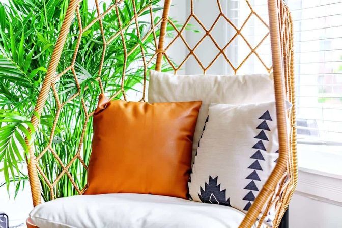 Sunlit indoor rattan hanging chair with white cushion, warm orange leather throw pillow and black-and-white geometric pillow, tucked beside a lush potted palm by a bright window — cozy boho reading nook.