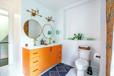 Bright modern bathroom with orange double vanity, round gold mirrors, brass fixtures, exposed brick accent, white tile floor and recessed shelf with plants.