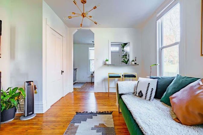 Sunny modern living room with green velvet sofa, textured throw and patterned pillows, hardwood floors, geometric rug, indoor plants, gold sputnik chandelier, large windows, and a small breakfast bar with two stools.