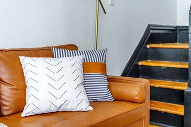 Stylish living room corner with a caramel leather sofa, white chevron and navy-striped throw pillows, brass floor lamp, and black staircase with wooden treads.