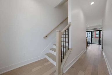 Bright modern apartment hallway with light hardwood floors, white walls, and a sleek wooden staircase with black metal balusters leading to balcony windows.