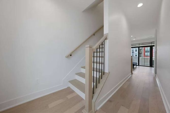 Bright modern apartment hallway with light hardwood floors, white walls, and a sleek wooden staircase with black metal balusters leading to balcony windows.
