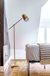 Cozy attic bedroom corner with brass adjustable floor lamp, white bed topped with a striped throw, natural jute rug and hardwood floor by a bright window